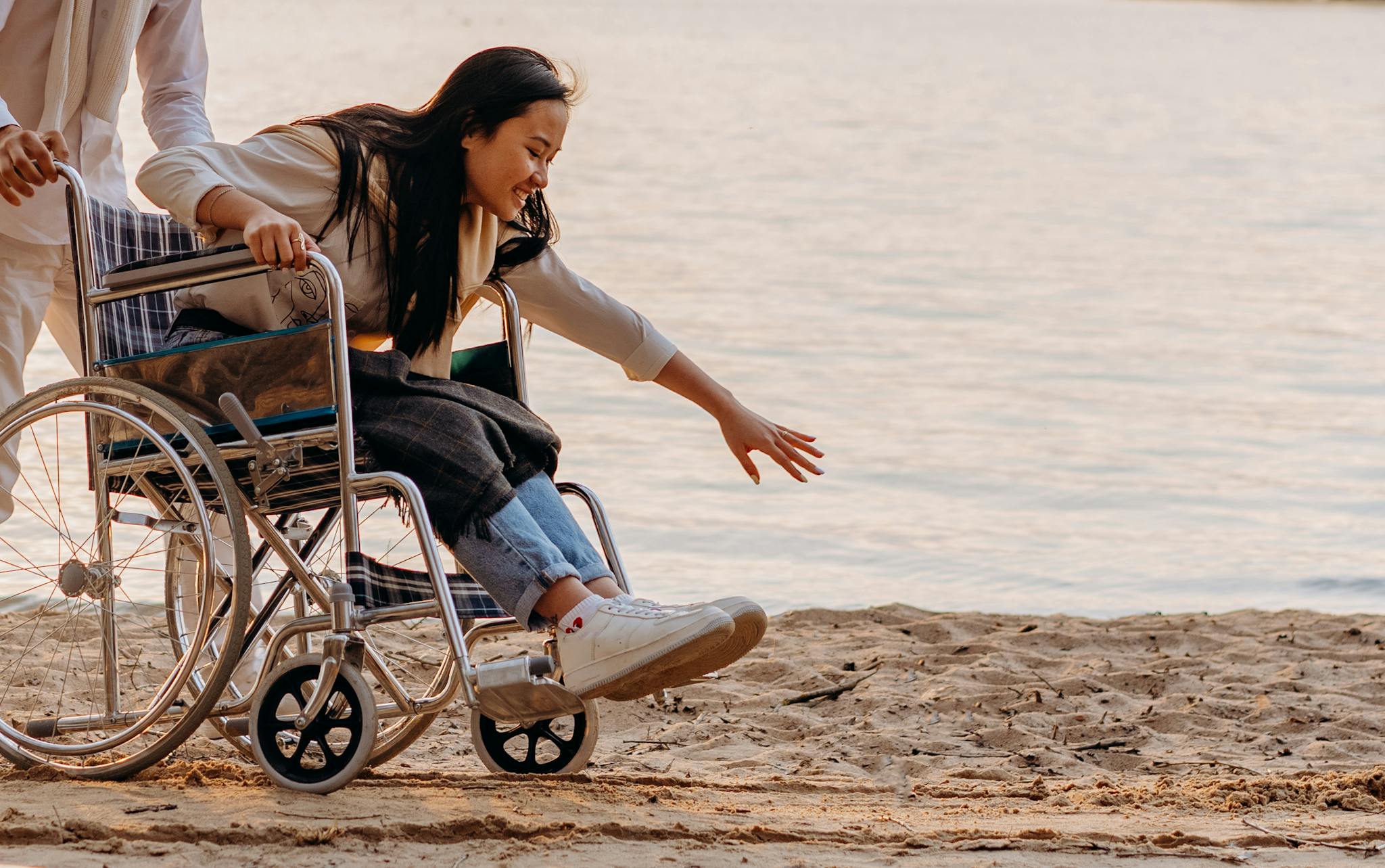Asian woman in a wheelchair enjoying time by the ocean on a sandy beach, reaching out.