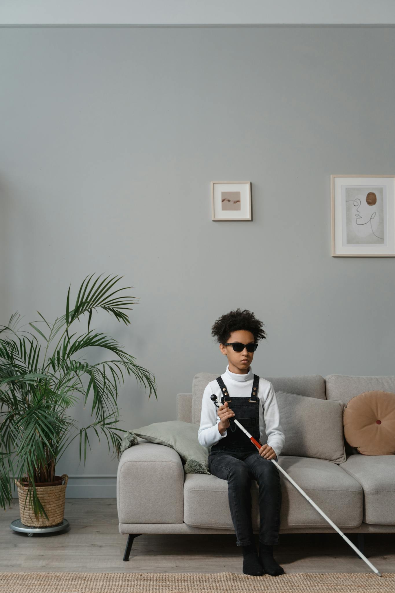 Young child with sunglasses holding white cane sitting on a sofa indoors.